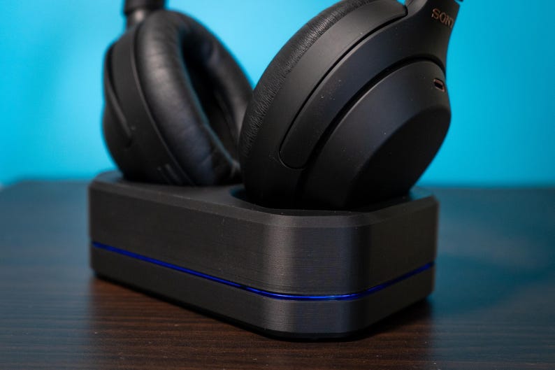 May include: Black over-ear headphones resting on a black charging dock with a blue accent line. The headphones are labeled "SONY" and have large, cushioned earcups. The dock is rectangular and sits on a dark wooden surface against a blue background.