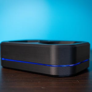 May include: A black rectangular device with a blue light strip. The device has a recessed top and sits on a dark wooden surface against a blue gradient background. The design is modern and minimalist.