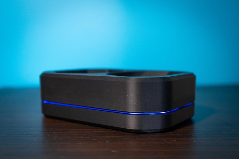 May include: A black rectangular device with a blue light strip. The device has a recessed top and sits on a dark wooden surface against a blue gradient background. The design is modern and minimalist.