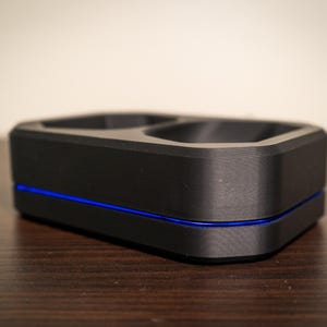 May include: A black, rectangular charging dock with two recessed areas. A thin blue line accents the base of the device. The dock is made of a matte material and sits on a dark wooden surface.
