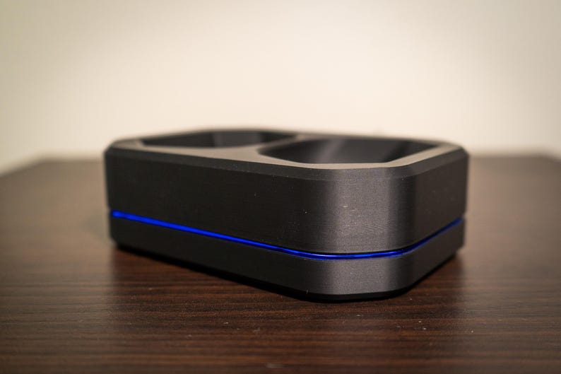 May include: A black, rectangular charging dock with two recessed areas. A thin blue line accents the base of the device. The dock is made of a matte material and sits on a dark wooden surface.