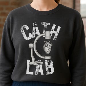 Puede incluir: Sudadera gris oscuro con las palabras "CATH LAB" en blanco, junto con una imagen estilizada de un corazón y un dispositivo médico. El diseño está centrado en la parte delantera de la sudadera.