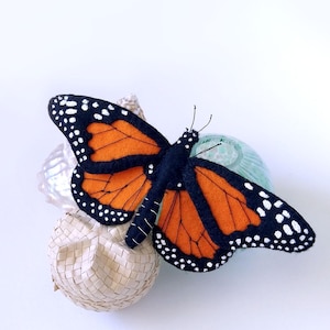 May include: A handcrafted monarch butterfly with orange and black wings, detailed with white spots. The butterfly is perched on a small, woven sphere and a light blue object, set against a white background. This decorative item is suitable for home decor.