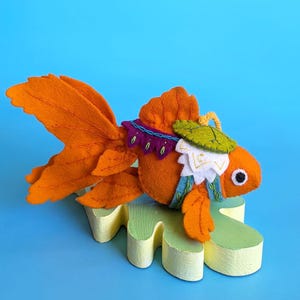 May include: A handcrafted orange felt goldfish ornament featuring a large flowing tail, a white and green saddle-like accessory with purple trim, and a black and white eye, resting on a pale yellow, leaf-shaped wooden base against a blue background.