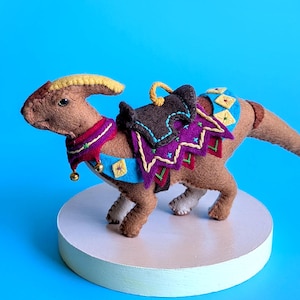 Può includere: Una statuetta di dinosauro in feltro marrone con una cresta gialla, che indossa una sella e un collare decorativi con motivi ricamati viola, blu e gialli e piccoli campanellini dorati sul collo, posizionata su una base rotonda bianca.