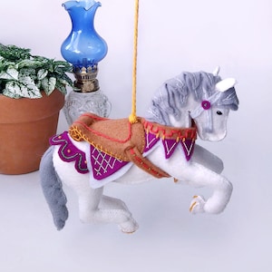 Può includere: Un ornamento a forma di cavallo da giostra in feltro bianco con criniera e coda grigie e una sella marrone e viola. Il cavallo è sospeso da uno spago giallo. Una lampada di vetro blu e una pianta in vaso sono sullo sfondo.