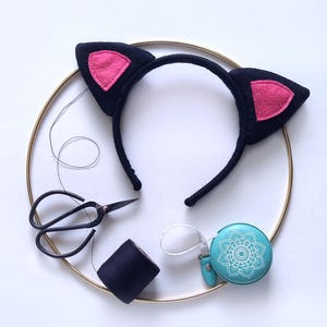 Puede incluir: Una diadema negra con orejas de gato y orejas internas rosas, una bobina de hilo azul oscuro, unas tijeras negras pequeñas y una cinta métrica turquesa con un diseño floral blanco, dispuestas sobre un aro dorado.