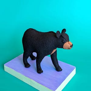 以下が含まれることがあります： フェルト素材で作られた手作りの黒いクマの置物で、日焼けした色の鼻と小さな丸い耳が特徴です。クマは4本の足で立ち、体の周りに目に見える縫い目があり、長方形の台座の上に配置されています。