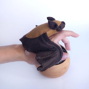 Può includere: Un giocattolo a forma di pipistrello fatto a mano in feltro marrone e nero. Il pipistrello ha grandi ali e piccole orecchie. Il giocattolo è appoggiato su una ciotola di legno. È un oggetto carino e stravagante.