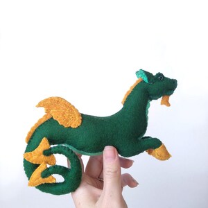 May include: A handmade felt dragon toy in shades of green, orange, and yellow. The dragon has a long, curved tail, wings, and small legs. The dragon is approximately 8 inches long.
