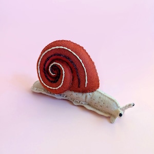 May include: A handcrafted felt snail featuring a rust-colored shell with a dark purple and white spiral stitch pattern. The snail body is a light beige felt with small brown speckles, two small antennae, and two black bead eyes on the front.