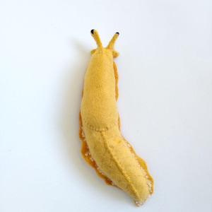 May include: A handmade, yellow felt slug with a stitched body and orange underside. The slug has two black-tipped antennae and a white, textured edge, creating a whimsical, soft sculpture. This unique item is perfect for home decor or as a playful gift.