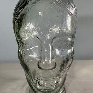Puede incluir: Una escultura de cabeza de vidrio transparente con un rostro detallado y una superficie texturizada. La cabeza tiene una línea de cabello definida y una base redonda. El vidrio tiene un ligero tinte verde y la escultura se exhibe sobre una superficie clara.