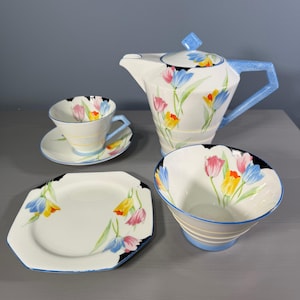 May include: A vintage tea set comprising a teapot, teacup, saucer, plate, and bowl. The white porcelain is adorned with tulip motifs in pink, yellow, and blue. The teapot features a blue handle and lid detail.