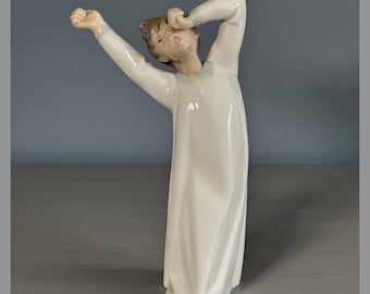 May include: A porcelain figurine of a child in a white nightshirt, stretching and yawning. The figurine is in a standing position with one arm raised above the head.