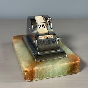 Peut inclure: Un calendrier perpétuel de bureau chromé vintage affichant "24 novembre". Le calendrier est posé sur une base rectangulaire en marbre vert et marron. Le calendrier est en métal brillant.