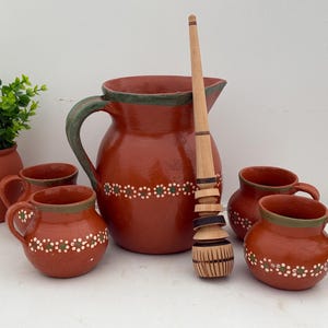 May include: A terracotta-colored pitcher, mugs, and a wooden whisk. The pitcher and mugs have a decorative white and green dotted pattern. The pitcher has a green handle and rim. The whisk is made of wood and has a carved design.