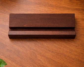 May include: A rectangular dark brown wooden stand featuring a horizontal groove carved across the center. The stand has a smooth, stained finish and a solid construction, designed to hold items upright in the slot while resting on a flat surface.