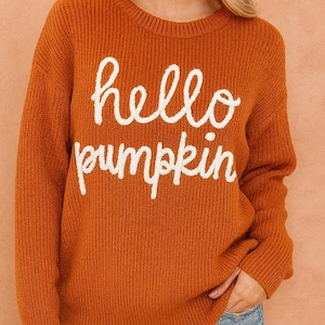 May include: An orange sweater with the words "hello pumpkin" in white script. The sweater has a ribbed knit texture and long sleeves. The model is wearing denim shorts.