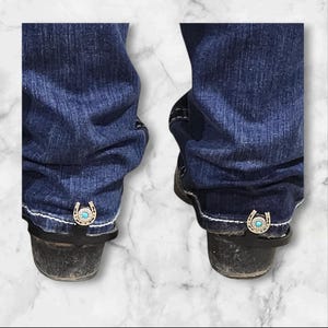 May include: A pair of blue jeans with a turquoise horseshoe charm attached to the bottom of each leg. The charms are attached to the jeans with a black strap.