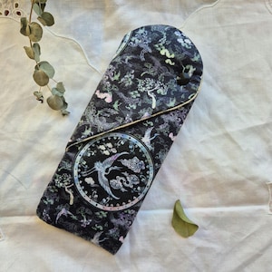 May include: A black fabric pouch with a floral and bird design. The pouch has a cream-colored trim and a flap closure.