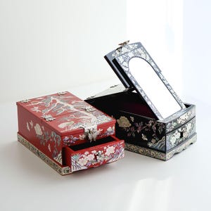 Puede incluir: Dos cajas de joyería decorativas con intrincados diseños florales y de pájaros. La caja roja tiene un cajón y la caja negra tiene un espejo y un cajón. Ambas cajas están hechas de madera e incrustadas con nácar.