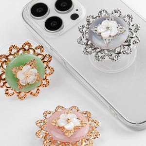 May include: Close-up of three decorative phone grips in gold, silver, and bronze tones. Each grip features a central, circular design in shades of green, pink, and grey, adorned with a white flower and ornate metalwork. The grips are attached to a clear phone case.