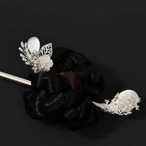 May include: A silver hair stick with ornate floral and leaf details, and a large, round, white pearl-like embellishment. The hair accessory is set against a dark background, showcasing its intricate design and elegant style.