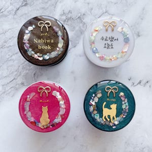 May include: Four round phone grips with decorative designs. One is brown with the text "Nabiwa book", another is white with Korean text, a third is pink with a gold cat, and the last is teal with a gold cat.