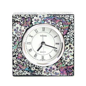 May include: Square quartz clock with a white face and black Roman numerals. The clock is surrounded by a black frame with an ornate design featuring birds and floral patterns in iridescent colours. The clock face reads "YUSUNG QUARTZ".