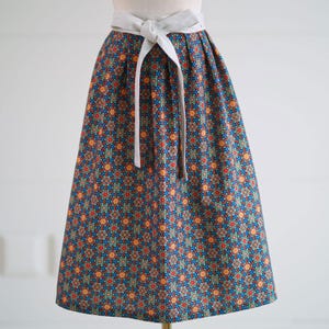 Puede incluir: Falda hasta la rodilla con fondo azul y un estampado floral repetido en rojo, naranja y turquesa. La falda tiene una cinturilla blanca y una cinta a juego. La falda está fruncida en la cintura y se ensancha.