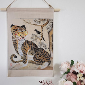 May include: A wall hanging featuring a traditional Korean tiger illustration. The tiger is depicted with orange and black stripes, yellow eyes, and a white chest. A black bird perches in a tree beside the tiger. The artwork is mounted on a beige fabric with a wooden rod.