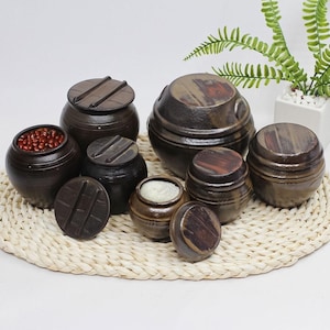 以下が含まれることがあります： 蓋付きの濃い茶色の陶器の瓶のコレクション。一部は開いており、豆や塩などの内容物が確認できます。瓶はサイズと形状が異なり、素朴で手作りの外観です。織りマットの上に配置されています。