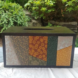 May include: A rectangular tissue box with a dark brown wooden frame. The front features panels of patterned fabric in gold, green, and rust. The top has an oval opening for tissue dispensing. The box is likely to be 25 cm long.