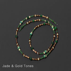 K&ouml;nnte beinhalten: Eine Halskette in Jade- und Goldt&ouml;nen. Die Halskette hat eine Kombination aus gr&uuml;nen, goldenen und braunen Perlen. Die Perlen sind zu einem langen, eleganten Design aufgereiht. Der Text "Jade & Gold Tones" steht unten.