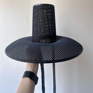K&ouml;nnte beinhalten: Schwarzer Hut mit breiter Krempe und zylindrischer Krone. Der Hut besteht aus einem Mesh-Material, und die Krempe hat eine leicht gebogene Form. Der Hut wird von einer Person gehalten, die eine schwarze Uhr tr&auml;gt.