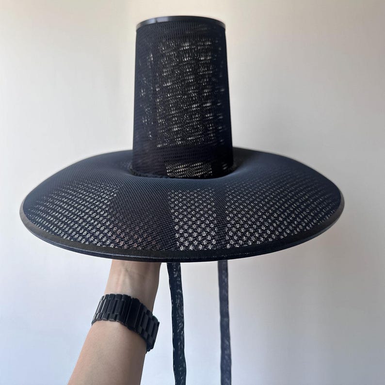 K&ouml;nnte beinhalten: Schwarzer Hut mit breiter Krempe und zylindrischer Krone. Der Hut besteht aus einem Mesh-Material, und die Krempe hat eine leicht gebogene Form. Der Hut wird von einer Person gehalten, die eine schwarze Uhr tr&auml;gt.