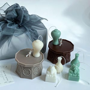 May include: A collection of decorative candles in different shapes and colours. Includes a white vase-shaped candle, a green vase-shaped candle, and a white candle with a detailed design. The candles are on a white surface.