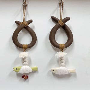 May include: Two hanging decorative ornaments. Each ornament features a brown, loop-shaped top with a jute twine accent. Below, a white tassel leads to a small, fish-shaped charm. One charm is light green, the other is white, both with small red accents.