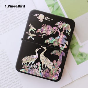 May include: A black lighter with a design of two cranes standing in front of a pine tree. The design is made of iridescent mother-of-pearl inlay.