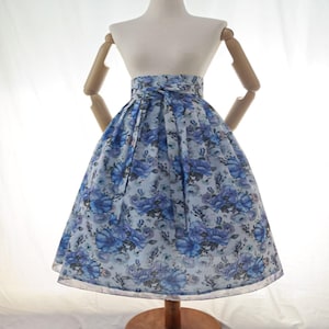 Puede incluir: Falda floral azul claro con cintura alta y lazo. La falda presenta un estampado de grandes rosas azules sobre un fondo azul claro. La falda se exhibe en un maniquí.