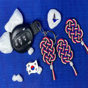 May include: A black car key fob with a silver keyring, accompanied by three red, white, and blue knot-shaped keychains. Also present are a small stone with a blue bird design and a stone with a South Korean flag.