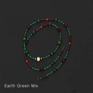 K&ouml;nnte beinhalten: Halskette Earth Green Mix mit gr&uuml;nen, braunen und orangefarbenen Perlen. Die Halskette hat eine Mischung aus runden und zylindrischen Perlen, mit einer cremefarbenen Perle in der Mitte. Der Text "Earth Green Mix" steht unten.