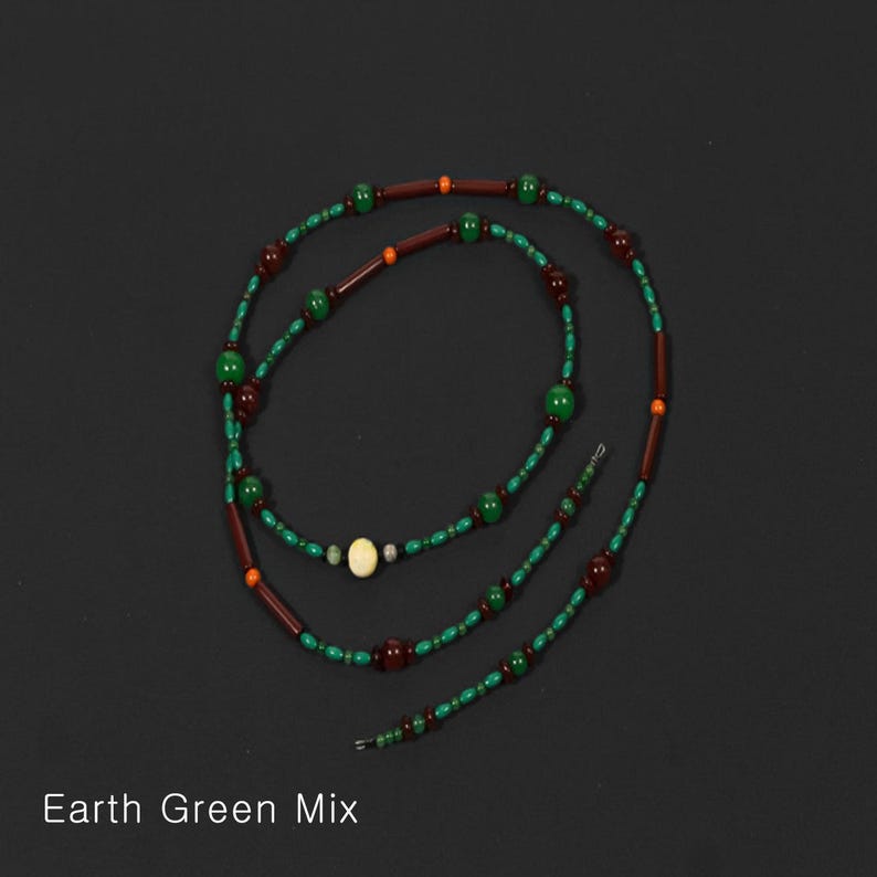 K&ouml;nnte beinhalten: Halskette Earth Green Mix mit gr&uuml;nen, braunen und orangefarbenen Perlen. Die Halskette hat eine Mischung aus runden und zylindrischen Perlen, mit einer cremefarbenen Perle in der Mitte. Der Text "Earth Green Mix" steht unten.