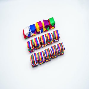 Op de afbeelding: Een assortiment kleurrijke, gevouwen stoffen ornamenten. De ornamenten zijn in rijen gerangschikt, met effen kleuren en verticale strepen in verschillende tinten. De levendige kleuren zijn rood, blauw, geel, groen en paars.