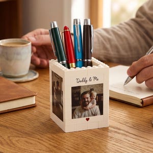 May include: A white pen holder made of interlocking blocks, featuring a photo and the text "Daddy & Me." It holds four pens in green, red, blue, and black. The holder sits on a wooden desk with a coffee cup and notebook.