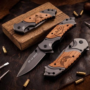 May include: Three folding pocket knives with grey metal frames and light brown wood handle inlays. One knife is open, displaying the date 07-12-2025 on the blade. The other two are closed, featuring engraved text Andrew, MD, and a winged emblem.
