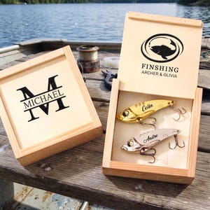 Könnte beinhalten: Geschenkbox aus Holz für Angelköder mit zwei Ködern im Inneren. Die Box ist geöffnet und zeigt einen goldenen und silbernen Köder. Der Deckel hat ein Angel-Logo und die Worte "FINISHING ARCHER & OLIVIA". Eine andere Box hat den Namen "MICHAEL" und den Buchstaben "M".