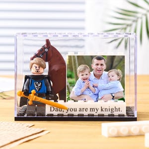 May include: A clear acrylic photo frame with a Lego-style base. Inside, a photo of a father and two children is displayed. A Lego knight figure and a brown horse are positioned in front of the photo. The base reads "Dad, you are my knight."
