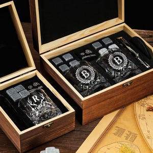 May include: Two wooden gift boxes with whiskey glasses, ice stones, and bar tools. One box has glasses with the letter "B" and a laurel wreath design. The other box has a glass with the letter "R" and the name "Robert". The boxes are on a map.